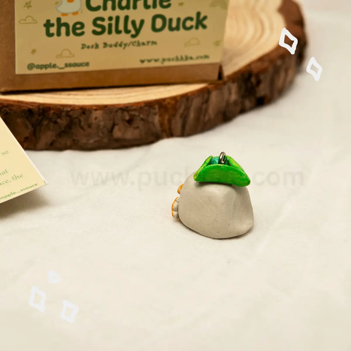 A small novelty desk buddy charm shaped like a duck sitting on a log, displayed on a white fabric surface with its branded cardboard box and a wood slice stand in the background.