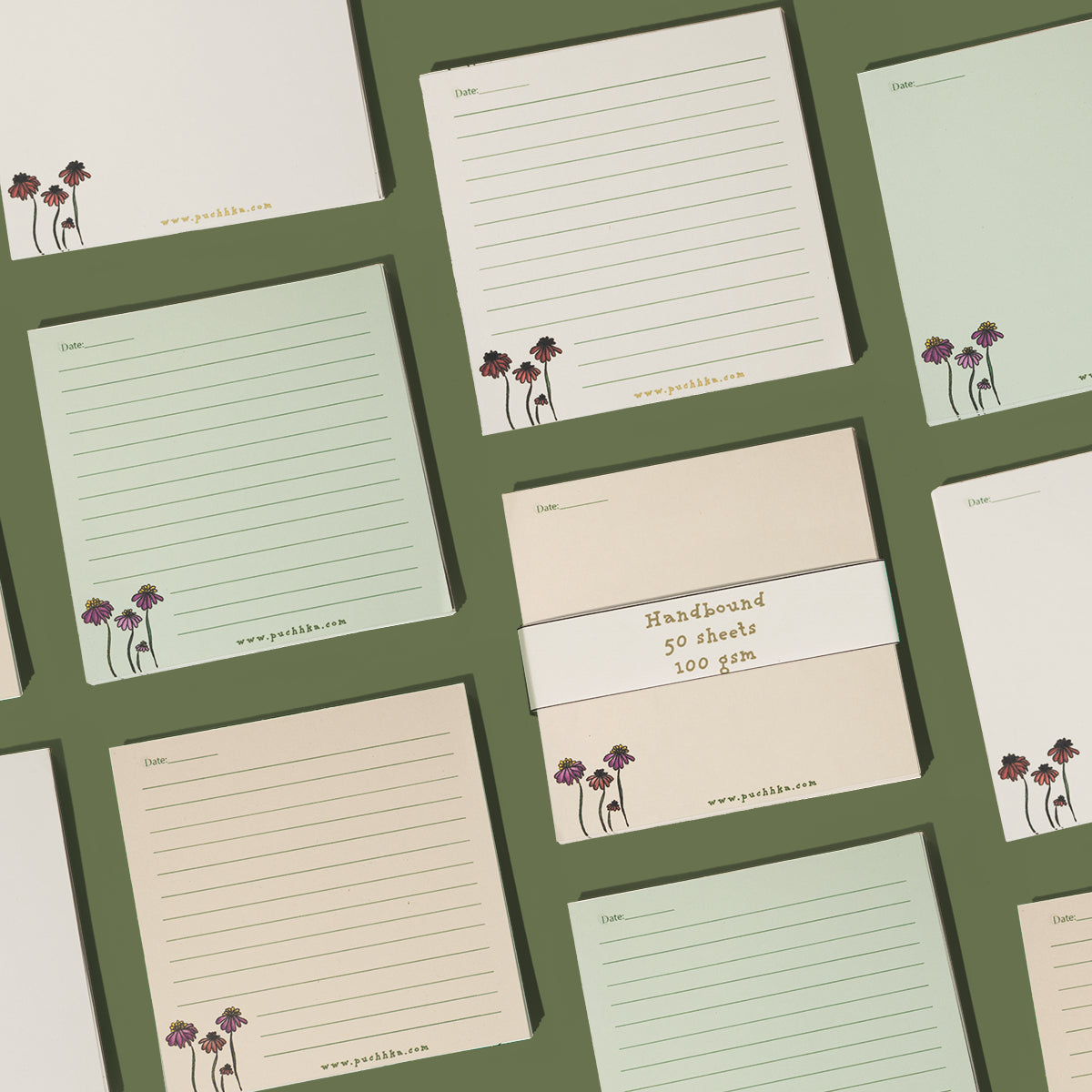 A set of lined notepads and a pack of unlined paper pads arranged in a flat-lay grid on a dark olive green background. The pads feature cream, light sage green, and pale peach colors, decorated with a small purple and gold floral illustration in the bottom-left corner 