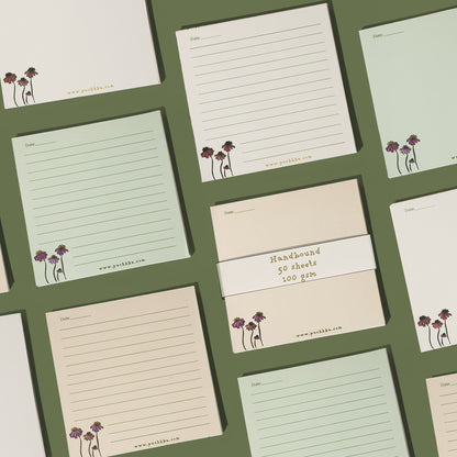 A set of lined notepads and a pack of unlined paper pads arranged in a flat-lay grid on a dark olive green background. The pads feature cream, light sage green, and pale peach colors, decorated with a small purple and gold floral illustration in the bottom-left corner 