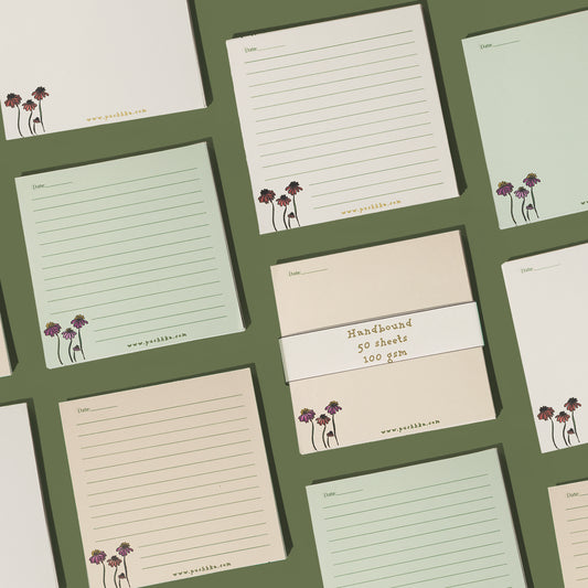 A set of lined notepads and a pack of unlined paper pads arranged in a flat-lay grid on a dark olive green background. The pads feature cream, light sage green, and pale peach colors, decorated with a small purple and gold floral illustration in the bottom-left corner 