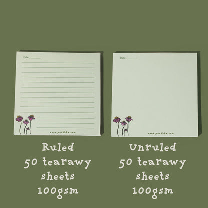 Two ConeFlowers Memopad notepads shown on a muted olive green background, labeled as ruled and unruled, each with 50 tearaway sheets, 100gsm. The notepads are off-white with a small purple and green flower illustration in the bottom-left corner.