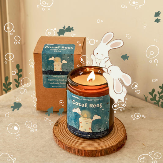 A lit amber glass jar candle labeled 'Coral Reef' sits on a round wood  coaster in front of its matching brown kraft box. The scene is styled with a warm beige background, whimsical white line-art of a rabbit and bubbles, and green leaf accents.