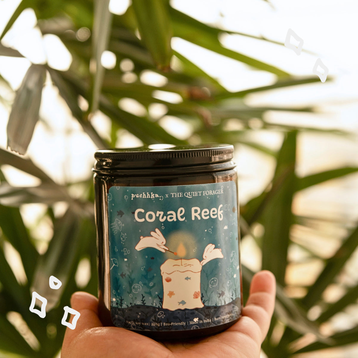 A hand holds a dark glass jar candle labeled 'Coral Reef', set against a blurred background of green palm leaves and warm sunlight.