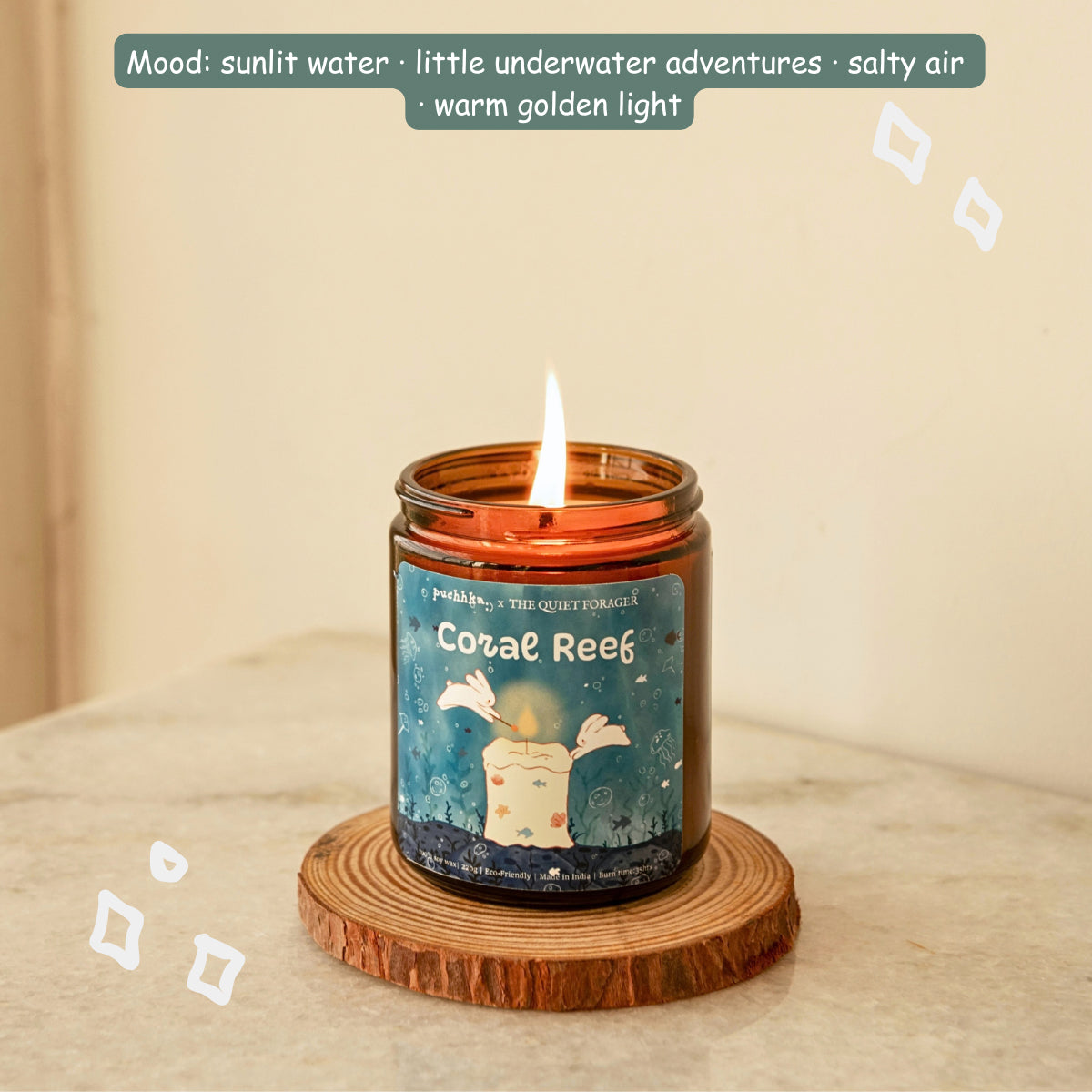 A lit amber glass jar candle labeled 'Coral Reef' sits on a round wooden coaster on a light stone surface, with a soft beige wall behind it and decorative text at the top describing the mood and scent.