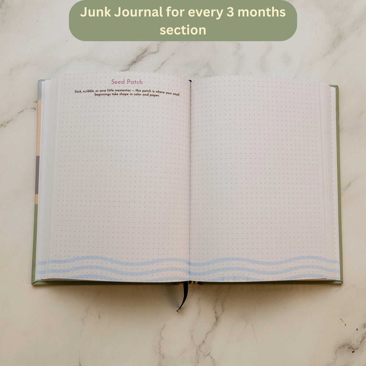 An open notebook journal on a light marble surface, showing blank lined pages with a green cover edge and a small ribbon bookmark. A green text bubble at the top reads 'Junk Journal for every 3 months section'.