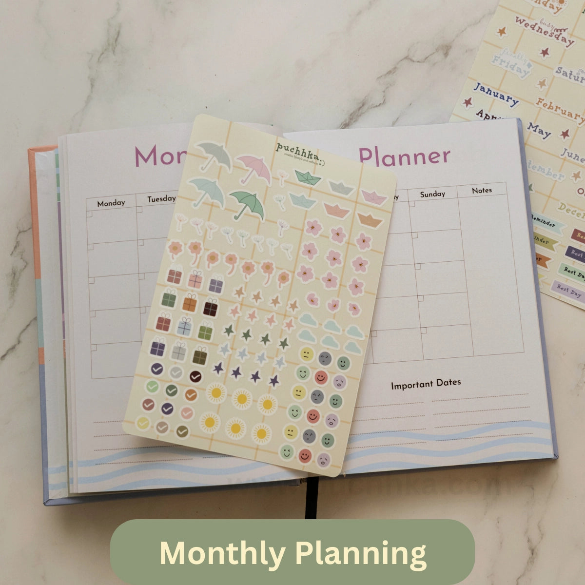 A monthly planner page spread open on a marble surface, with a sheet of decorative planner stickers placed on top. The stickers include pastel umbrellas, stars, and date icons.