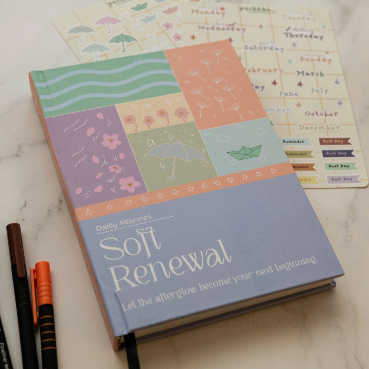 A hardcover daily planner titled 'Soft Renewal' lies on a light marble surface, showing pastel illustrated interior pages and a lavender cover. Two pens are placed beside it, and part of a matching sticker sheet is visible in the background.
