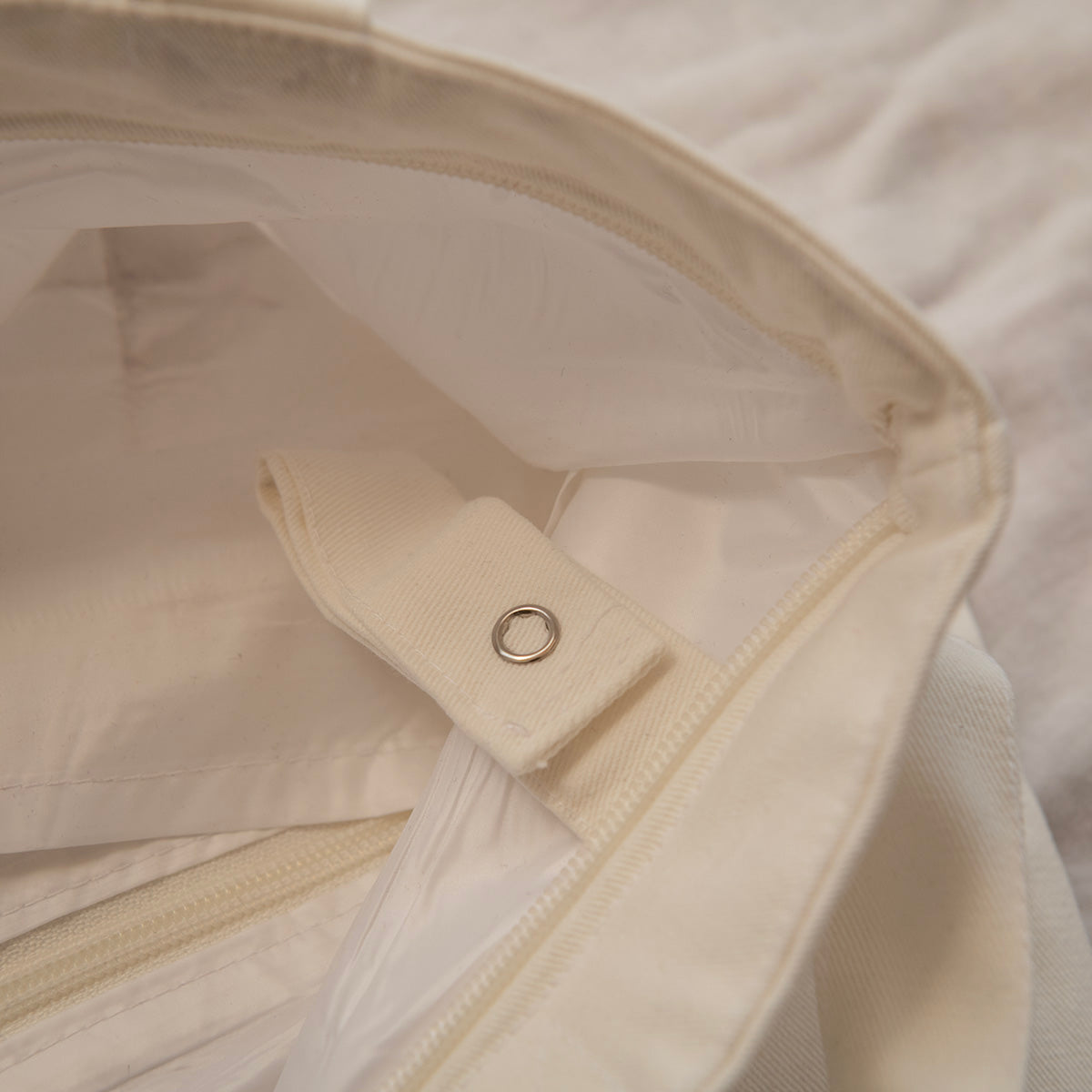 Close-up detail of a cream-colored canvas bag showing the interior lining, zipper track, and a fabric tab with a metal grommet.