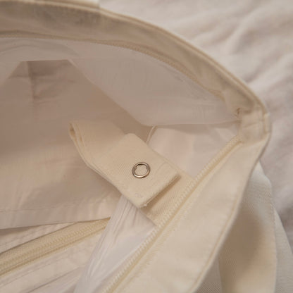 Close-up detail of a cream-colored canvas bag showing the interior lining, zipper track, and a fabric tab with a metal grommet.