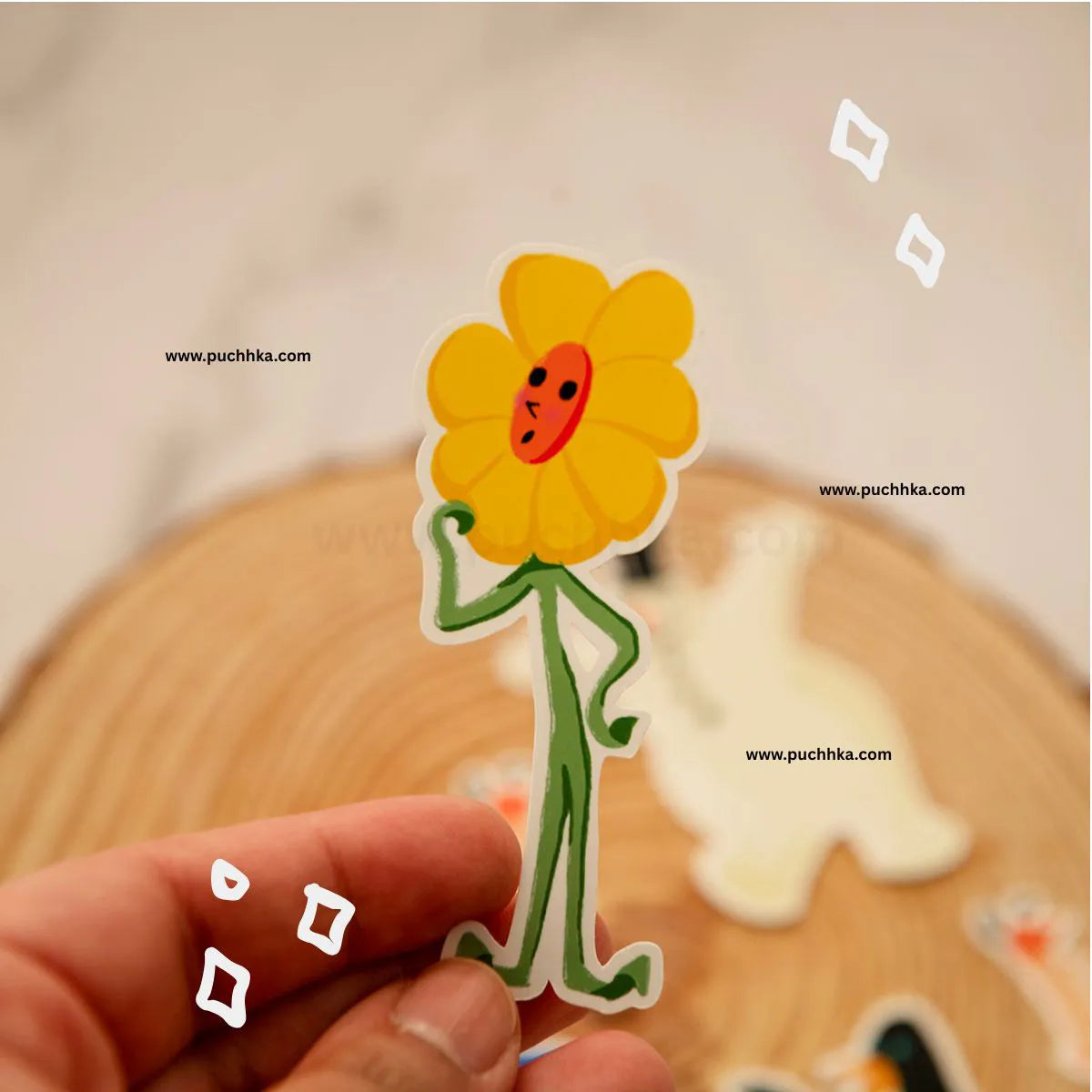 A hand holds a die-cut sticker of a smiling yellow flower with green limbs and a red face, with other blurred stickers and a wooden surface in the background. 