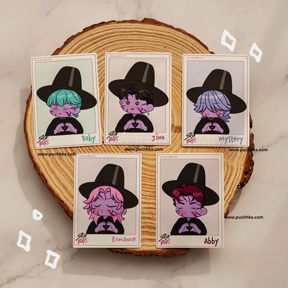 Five illustrated polaroid-style cards featuring cartoon characters wearing black hats are arranged on a round wood slice coaster against a light marble surface. The cards are labeled Baby, Jinu, Mystery, Romance, and Abby, with the brand name Puchhka visible in small text.
