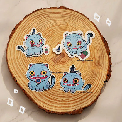 Four cartoon cat stickers in light blue with colorful expressions are arranged on a round wood slice background. The stickers have white borders and small printed website text, photographed on a light marble-like surface.