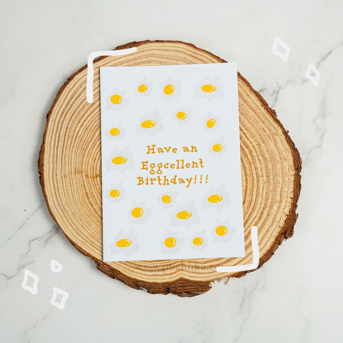 A white greeting card with a playful egg-themed design and the text 'Have an Eggcellent Birthday!!!' in orange, styled on a round wood slice coaster over a light marble surface.