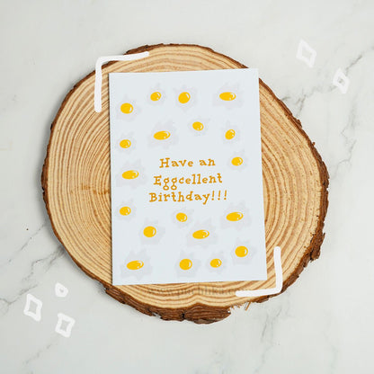 A white greeting card with a playful egg-themed design and the text 'Have an Eggcellent Birthday!!!' in orange, styled on a round wood slice coaster over a light marble surface.