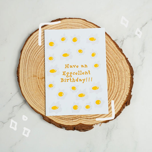 A white greeting card with a playful egg-themed design and the text 'Have an Eggcellent Birthday!!!' in orange, styled on a round wood slice coaster over a light marble surface.