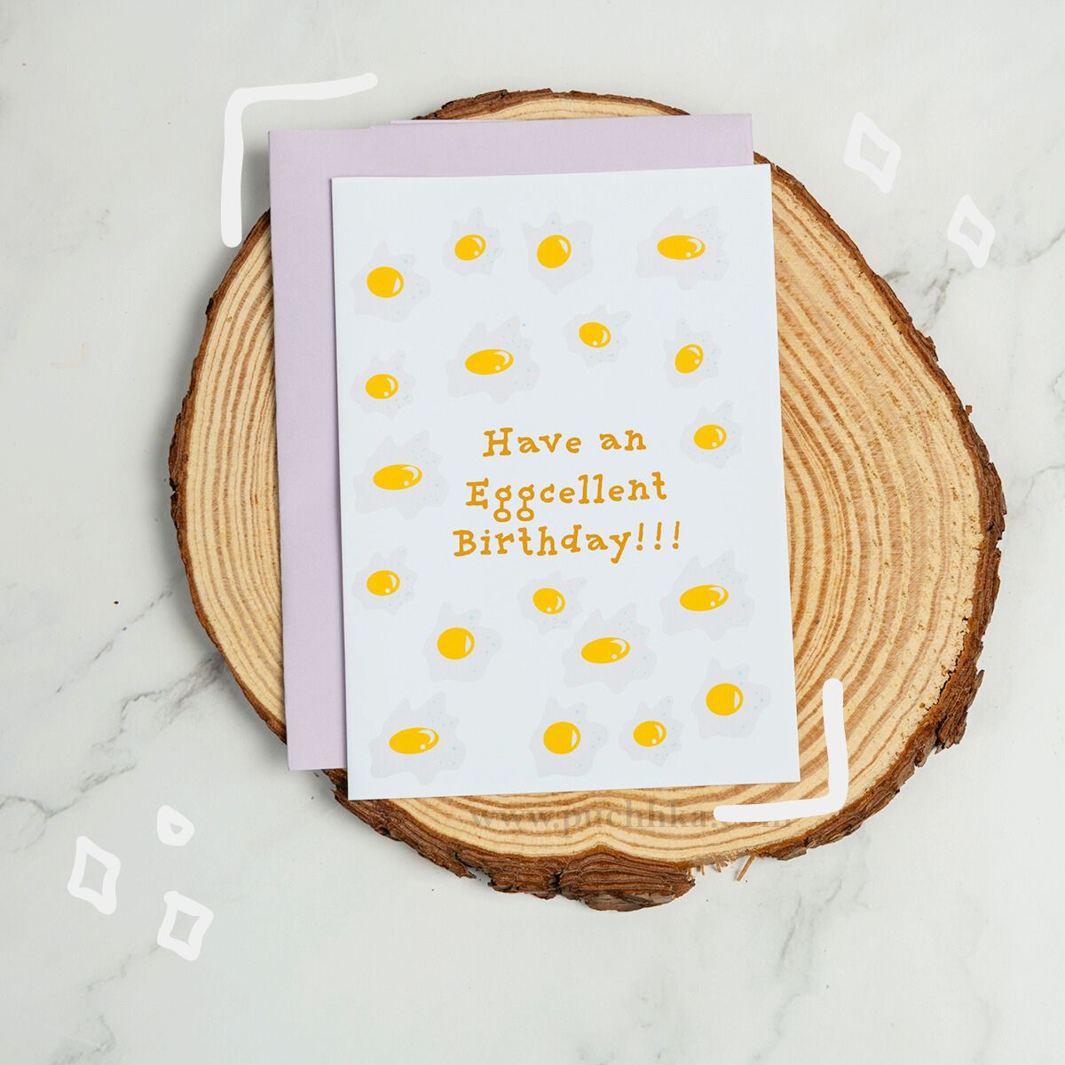 A greeting card with a white front and lavender envelope, placed on a round wood slice over a light marble surface. The card is printed with yellow egg yolk shapes and the text 'Have an Eggcellent Birthday!!!'.