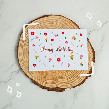 A square white birthday card with colorful floral illustrations and the words 'Happy Birthday' in cursive, placed on a round wood slice coaster over a light marble surface.