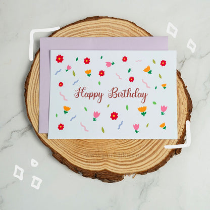A floral 'Happy Birthday' greeting card with a lavender envelope, displayed on a round wood slice coaster over a light marble surface.