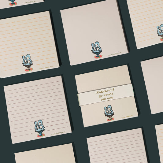 A flat-lay arrangement of cream-colored notepads with various line styles and a small fountain printed near the bottom. One central notepad is wrapped in a cream paper band labeled 'Handwritten 50 sheets 100 gsm'.