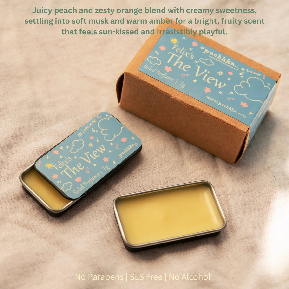 A small rectangular metal tin of solid perfume shown open and closed on a beige fabric surface, alongside its kraft paper box. The lid and box feature a light blue label with the scent name 'Felix's The View' and decorative cloud and flower illustrations.