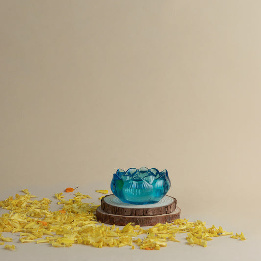 A small translucent blue lotus-shaped bowl or candle holder sits on stacked wood coaster, surrounded by scattered yellow flower petals against a plain beige background.