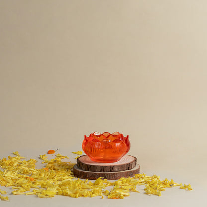 A translucent orange-red lotus-shaped bowl or candle holder displayed on two stacked wood slices, with scattered yellow flower petals on a beige surface against a plain beige background.
