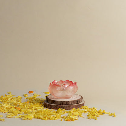 A translucent pink lotus-shaped bowl or candle holder displayed on two stacked wood slices, surrounded by scattered yellow flower petals against a warm beige background.