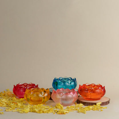 Five small lotus-shaped glass candle holders in red, orange, pink, yellow, and blue, arranged on a beige surface with scattered yellow petals and one on a wooden coaster.