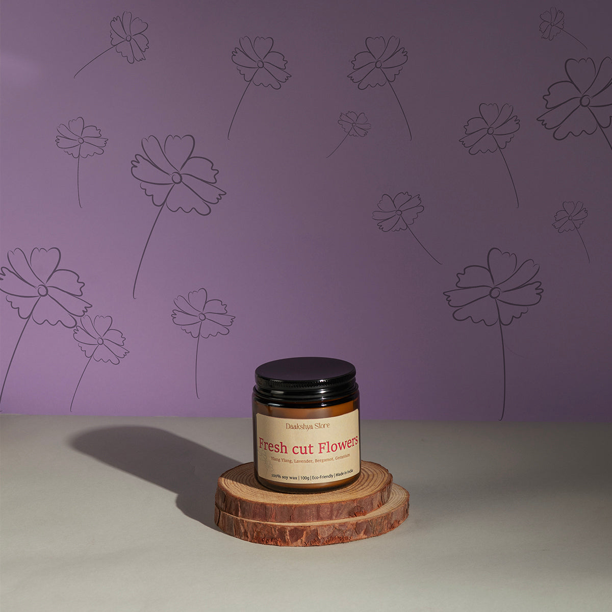 A jar candle labeled 'Fresh Cut Flowers' by Daakshya Store (now Puchhka) sits on two stacked wood coaster against a lavender wall with floral line drawings.
