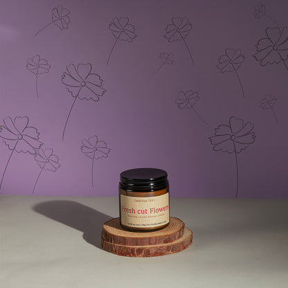 A jar candle labeled 'Fresh Cut Flowers' by Daakshya Store (now Puchhka) sits on two stacked wood coaster against a lavender wall with floral line drawings.