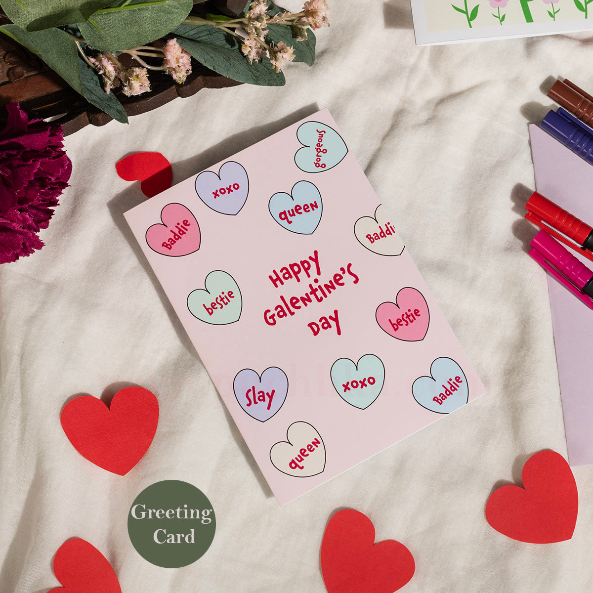 A pink Valentine's Day greeting card with colorful heart illustrations and the words 'Happy Galentine's Day' in red, styled on a cream fabric background with red paper hearts, flowers, and stationery.