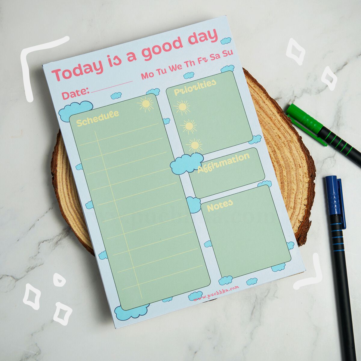A daily planner notepad titled 'Today is a good day' with pastel green writing sections, shown on a wood slice coaster with two pens nearby on a marble surface.