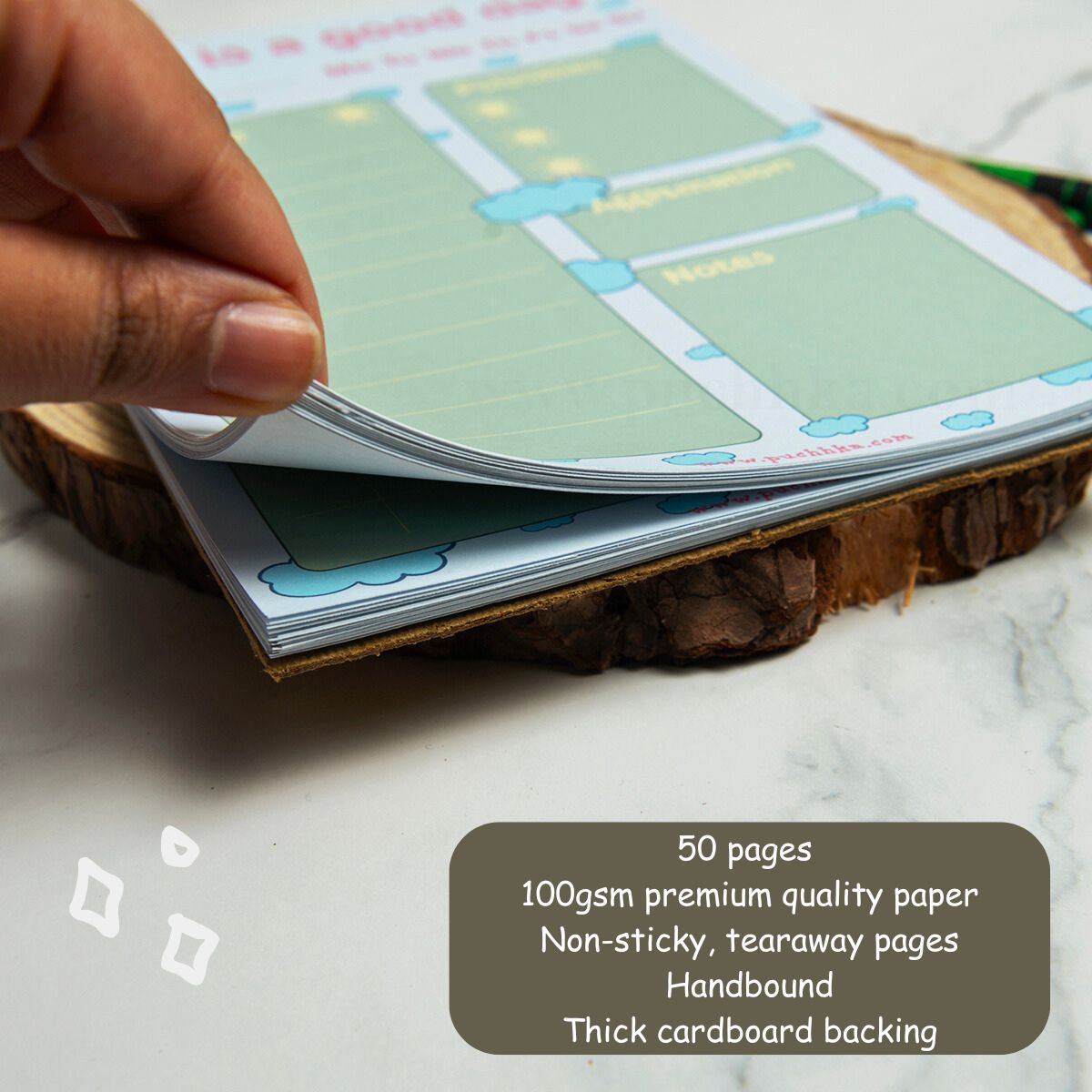 A hand is lifting a page from a tearaway notepad with a light green and blue planner-style layout, resting on a rustic wood slice. A dark gray text box in the lower right lists product details: 50 pages, 100gsm premium quality paper, non-sticky tearaway pages, handbound, and thick cardboard backing.