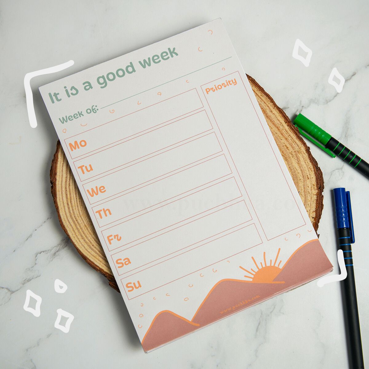 A weekly planner notepad titled 'It is a good week' with fields for Week of, Mo through Su, and a Priority column, styled on a wood slice coaster with pens nearby on a marble surface.