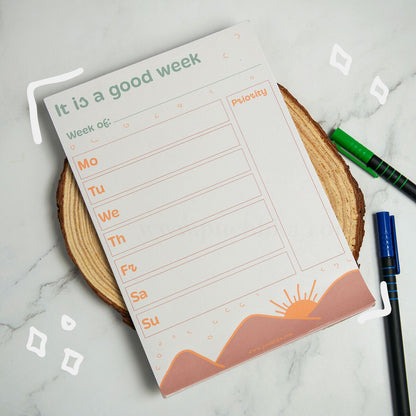 A weekly planner notepad titled 'It is a good week' with fields for Week of, Mo through Su, and a Priority column, styled on a wood slice coaster with pens nearby on a marble surface.