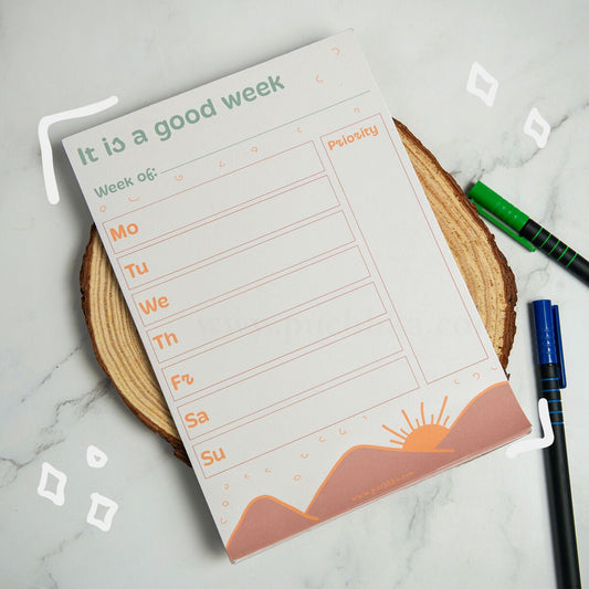 A weekly planner notepad titled 'It is a good week' with fields for Week of, Mo through Su, and a Priority column, styled on a wood slice coaster with pens nearby on a marble surface.