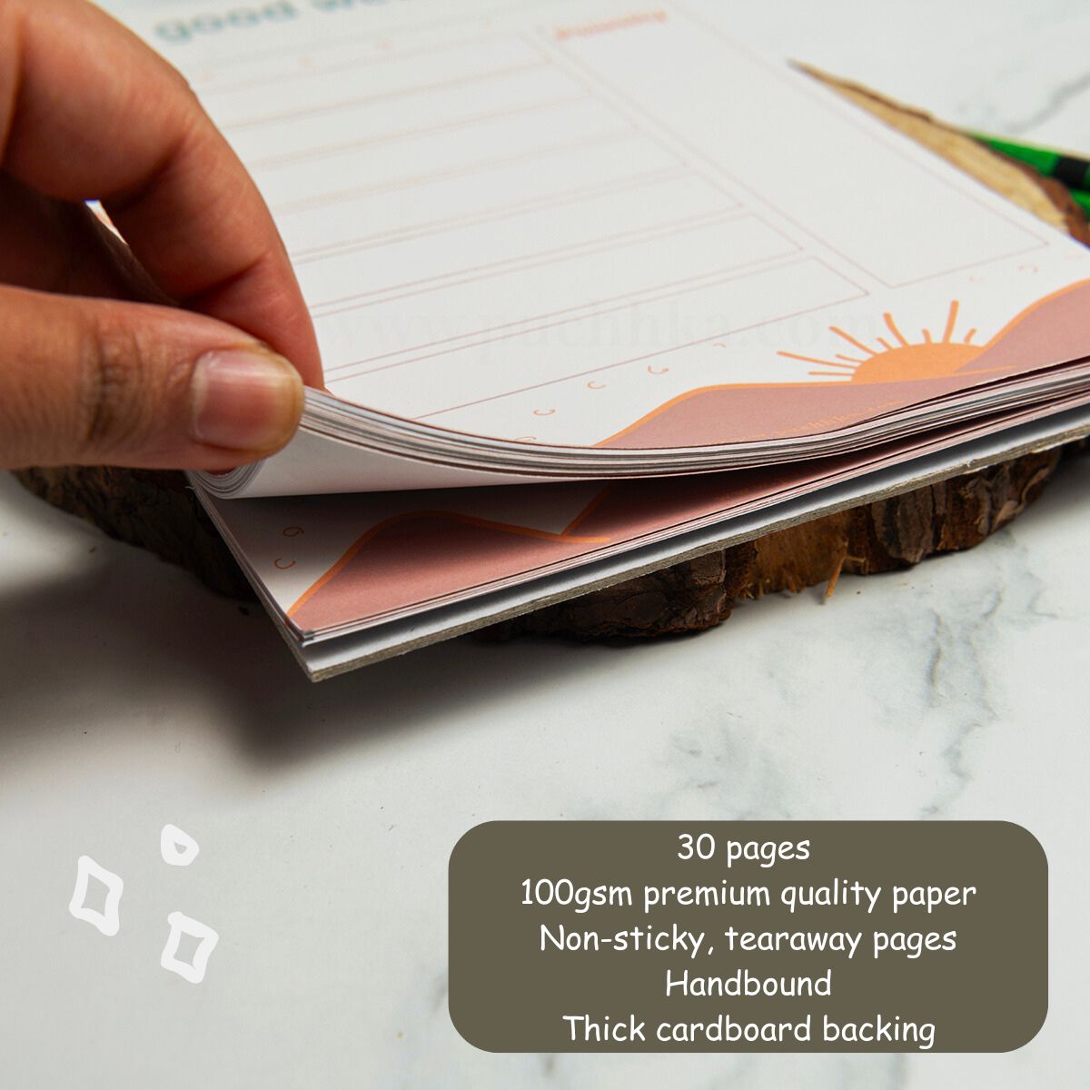 A close-up of a hand flipping through printed tearaway planner or notepad pages resting on a wood coaster. A dark text box in the lower right lists product details: 30 pages, 100gsm premium quality paper, non-sticky tearaway pages, handbound, and thick cardboard backing.