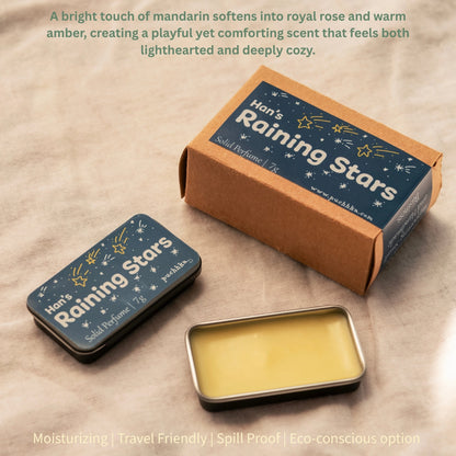 A small rectangular metal tin of solid perfume with pale yellow wax inside sits on a beige fabric surface, alongside its kraft paper box and a smaller matching tin. The labels read 'Han's Raining Stars' and 'Solid Perfume 7g'.