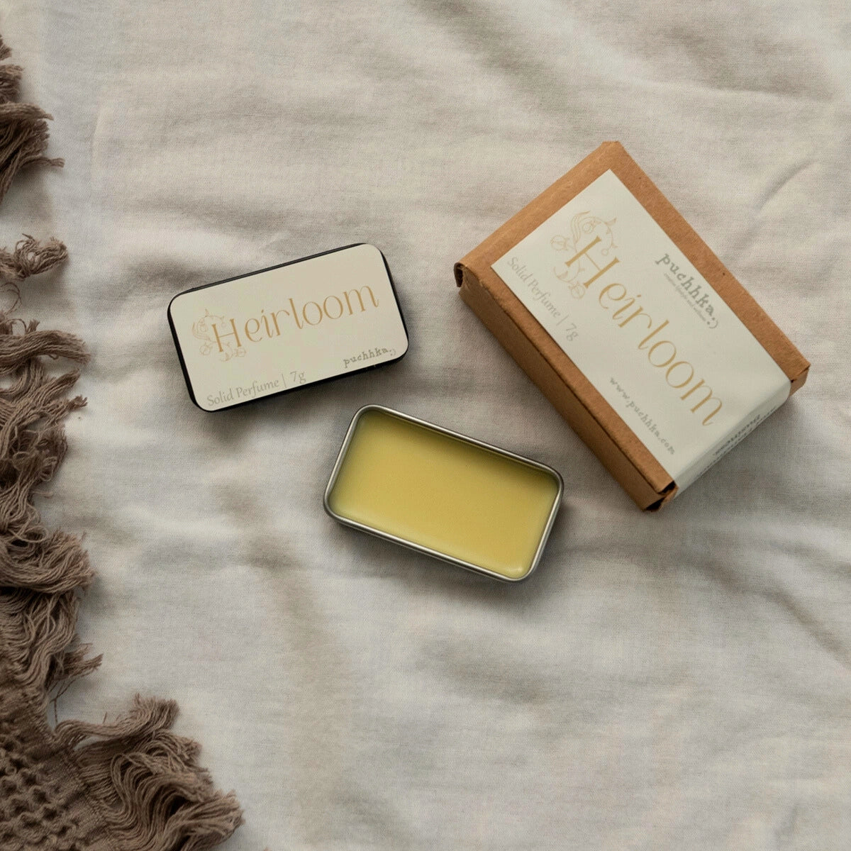 A flat-lay product photo of a 'Heirloom' Solid Perfume by Puchhka. The image shows an open silver tin filled with pale yellow perfume balm, a closed black tin with a cream label, and its brown kraft paper packaging box, all arranged on a textured beige linen fabric next to a brown fringed textile.