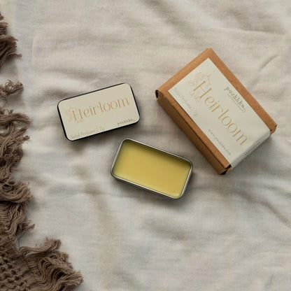 A flat-lay product photo of a 'Heirloom' Solid Perfume by Puchhka. The image shows an open silver tin filled with pale yellow perfume balm, a closed black tin with a cream label, and its brown kraft paper packaging box, all arranged on a textured beige linen fabric next to a brown fringed textile.