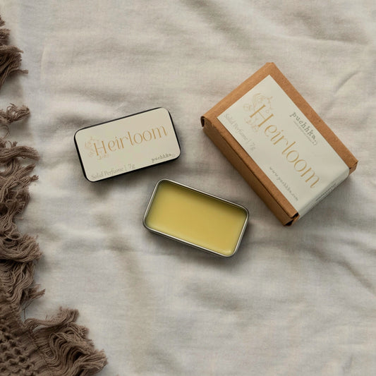 A flat-lay product photo of a 'Heirloom' Solid Perfume by Puchhka. The image shows an open silver tin filled with pale yellow perfume balm, a closed black tin with a cream label, and its brown kraft paper packaging box, all arranged on a textured beige linen fabric next to a brown fringed textile.