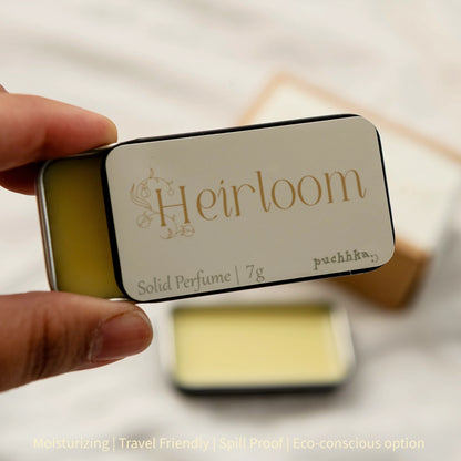 A hand holding a small rectangular metal tin of Heirloom solid perfume, with the lid facing the camera and showing the brand name. The background is softly blurred, showing another tin and a box.