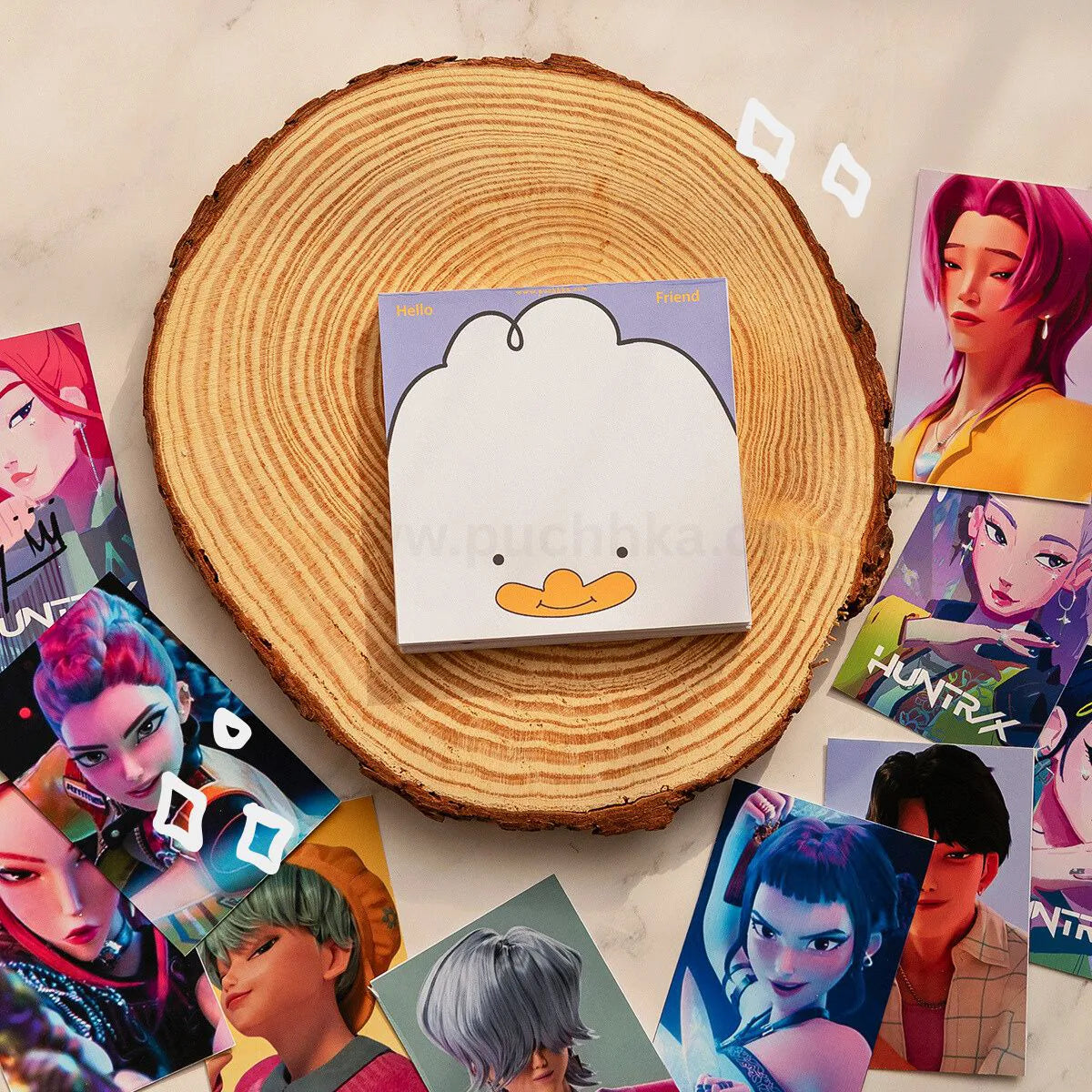 A square notepad with a cartoon duck face sits centered on a round wood slice, surrounded by scattered kpop demon hunters character cards on a light marble-like surface.