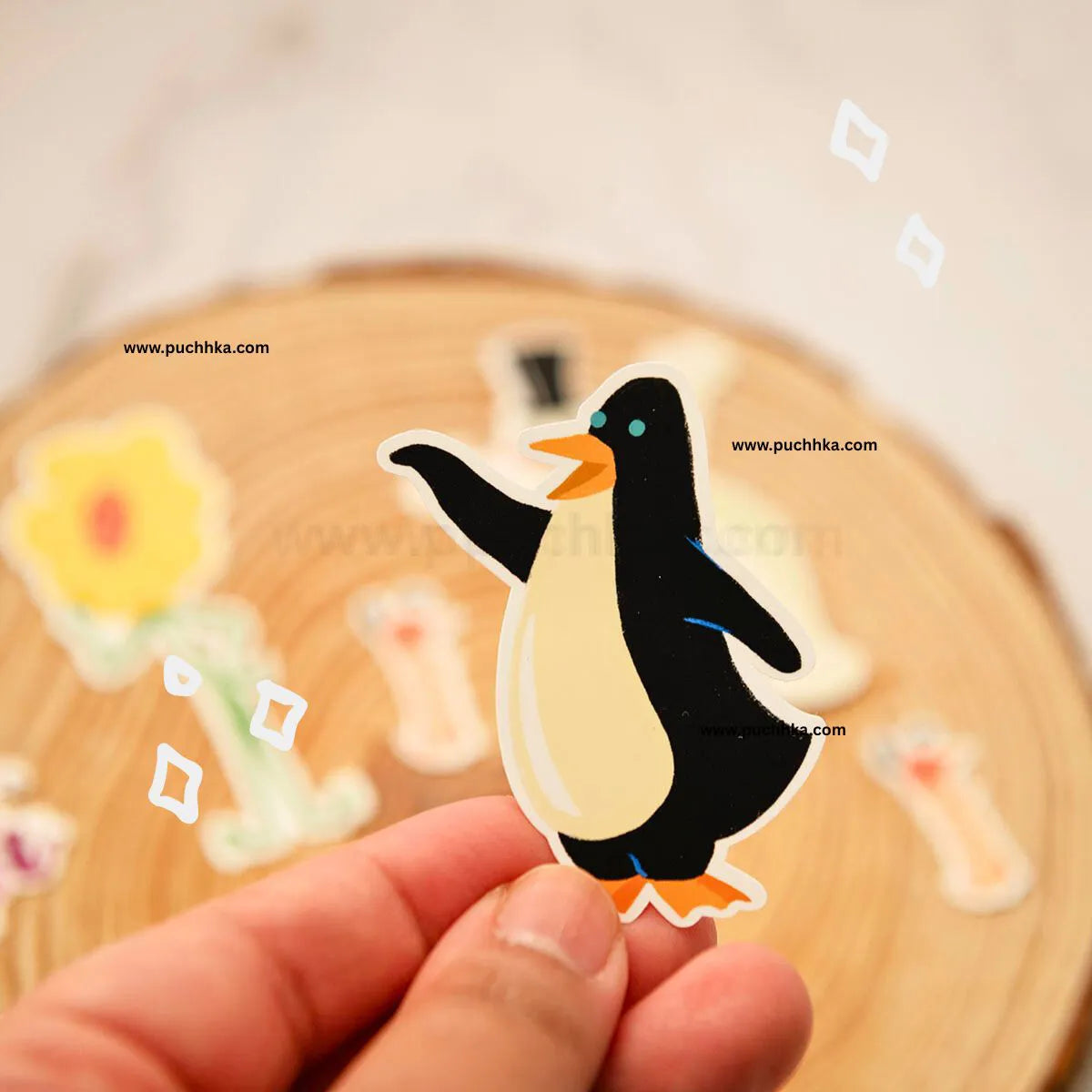 A hand holds a die-cut sticker of a penguin with a black body, cream belly, orange beak and feet, against a blurred wooden slice background with other stickers.