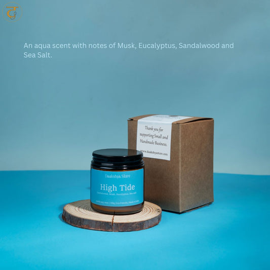 A jar of 'High Tide' scented candle with a black lid and blue label, displayed on a wooden  coaster next to its brown kraft box, set against a blue background with descriptive text overlay.