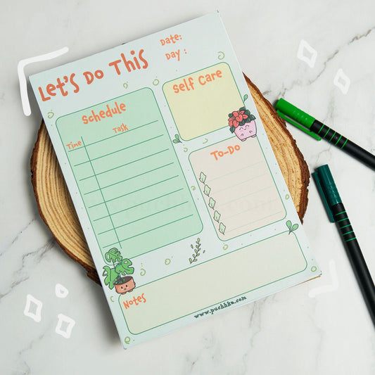 A pastel planner notepad titled 'Let's Do This' with sections for schedule, self care, to-do, and notes, styled on a wood slice with two pens on a marble surface.