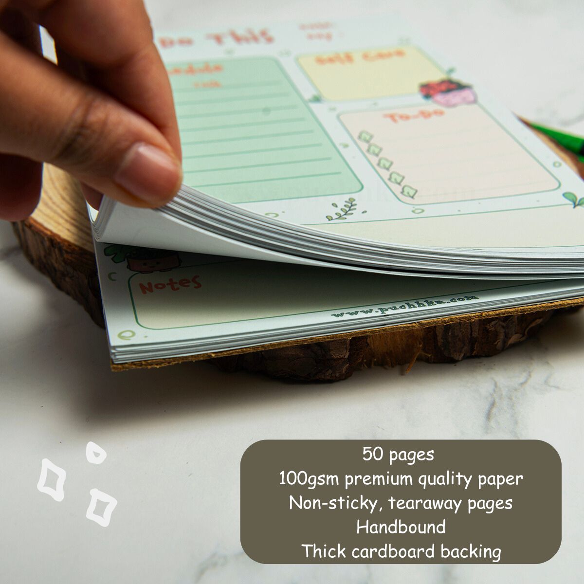 A close-up of a hand flipping through the pages of a planner notepad set on a rustic wood slice coaster. The notepad features pastel green and yellow sections with cute illustrations.