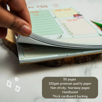 A close-up of a hand flipping through the pages of a planner notepad set on a rustic wood slice coaster. The notepad features pastel green and yellow sections with cute illustrations.