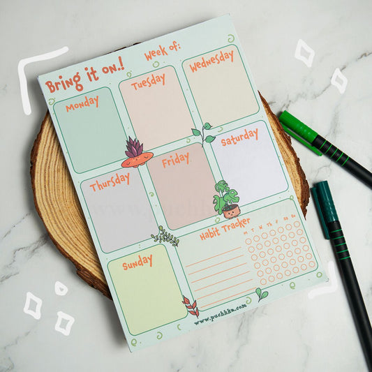 A weekly planner insert titled "Bring it on!" with sections for Monday through Sunday, a habit tracker, and small plant illustrations, styled on a wood slice with two green pens on a marble surface.