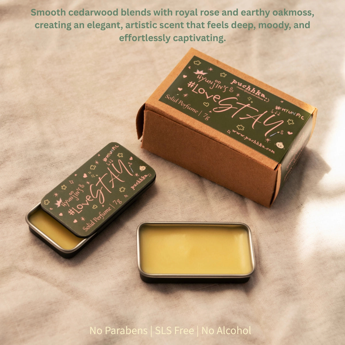 A small rectangular metal tin of balm with pale yellow wax sits in front of its kraft paper box and matching lid, all printed with the scent name "Hyunjin's Lovestay"and brand "Puchhka". The products are arranged on a beige fabric background with descriptive text at the top and bottom.